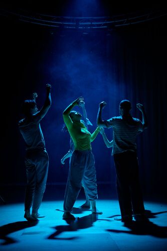 An ensemble of dancers with faces raised to the ceiling lit in blue on stage