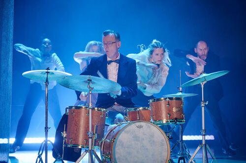Conor Guilfoyle seated at a set of drums with four dancers in movement behind