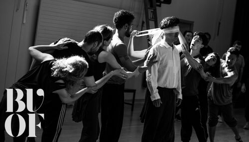 An ensemble gathered around a blindfolded figure in black and white