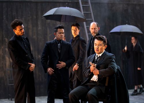 Cast members all in black looking towards a figure seated at the front of the frame