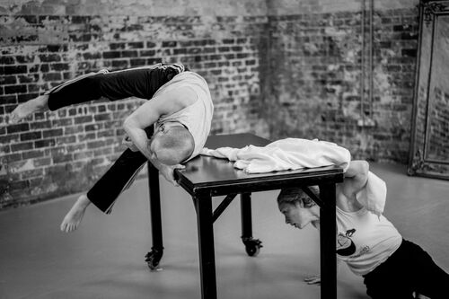 Jonathan Mitchell mid-roll over a table as Justine Cooper moves beneath it in black and white