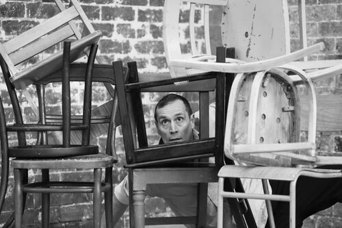 Lee Clayden in black and white looking through a wall of stacked chairs