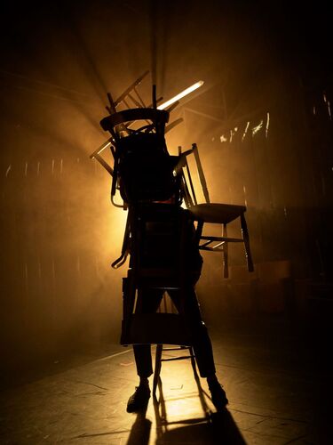 A dancer silhouetted carrying a mass of chairs on their body