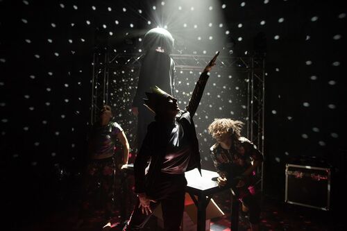 Three dancers in movement around a fourth figure standing on a table with a disco ball headpiece