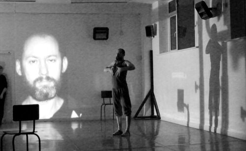 Jonathan Mitchell in movement in a studio with his face projected on the wall beyond