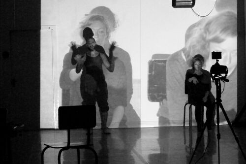 One figure standing and one seated in front of a camera with projections cast on both