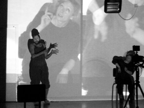 One figure standing and one seated in front of a camera with projections cast on both