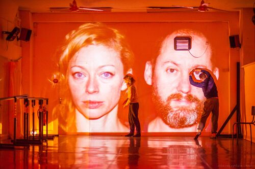 Two faces projected on the studio wall in orange with two figures in movement in front of them