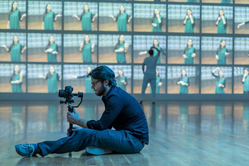 Christopher Ash sitting on the floor with a camera with a dancer moving in the background