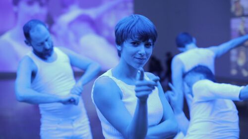 Ivonne Kalter lit in blue pointing and looking to the camera with others dancing behind