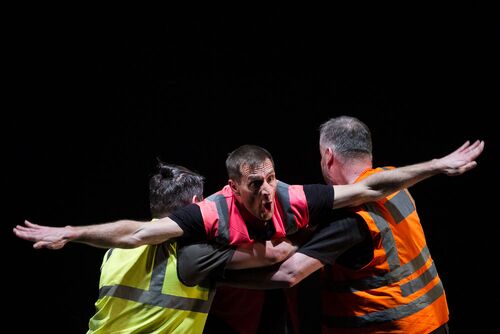 Two figures in hi-vis vests lifting a third figure with arms spread wide