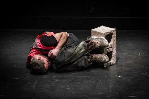 A figure in a pink hi-vis vest curled on the floor with a cinder block at his feet