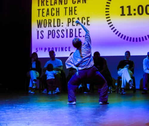 A dancer in movement seen from behind with judges seated in the background