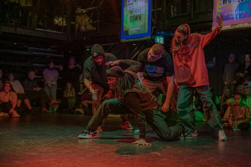 A dancer in movement on the floor with three others standing over him