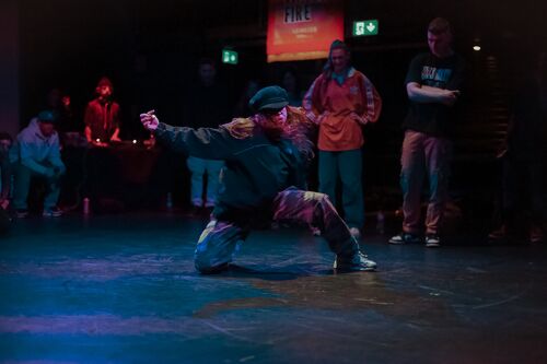 A dancer in movement on the floor with others gathered behind watching