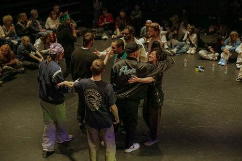 A group of dancers smiling and embracing in a black box space