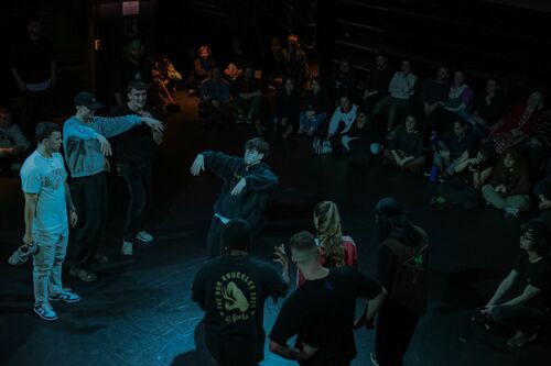 A figure in movement surrounded by other dancers standing and an audience seated behind