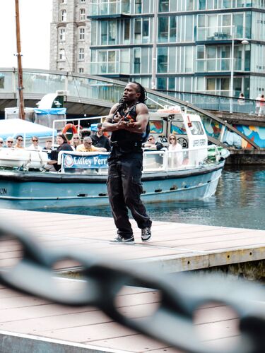 A figure standing on a dock with a boat on the water in the background