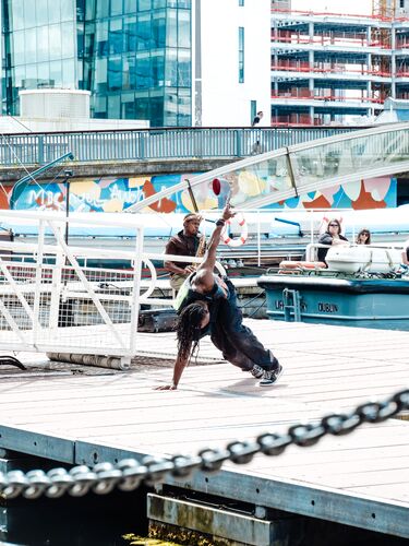 A figure in movement on a dock with another figure playing saxophone seen behind